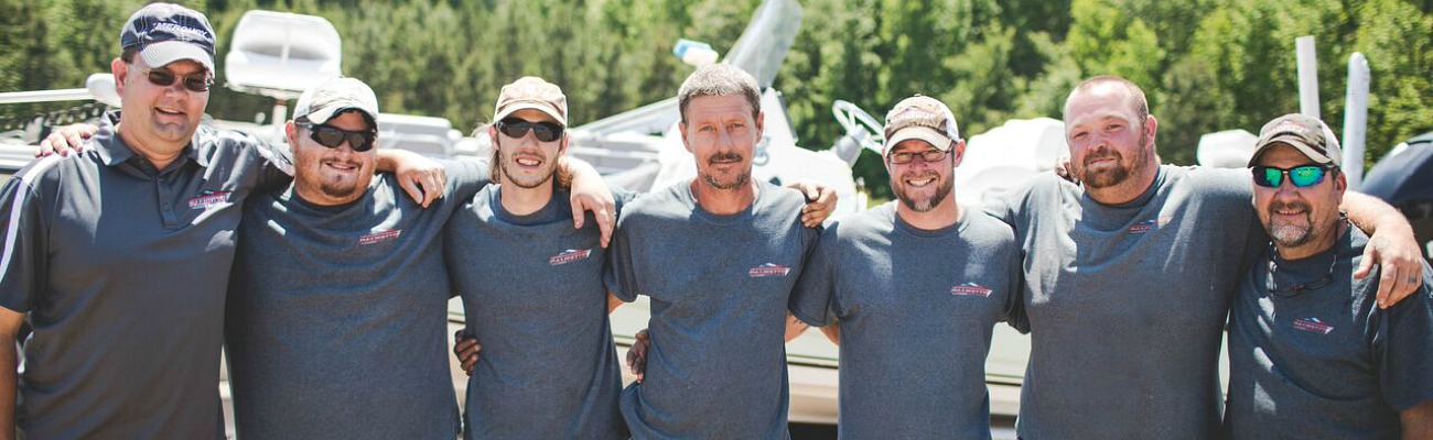 Seven people on the Palmetto Boat Center Service team standing side by side with arms around each other outside with boats and trees in background.